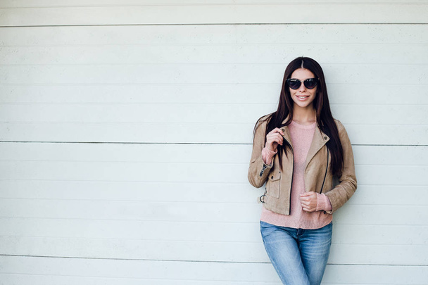 Candid and stylish beauty. Beautiful young woman in sunglasses adjusting her jacket and looking at camera with smile while standing outdoors - Photo, Image