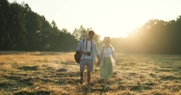 Young Caucasian happy girlfriend and boyfriend walking hand in hand with a camera in the field on the summer sunset. - Materiał filmowy, wideo