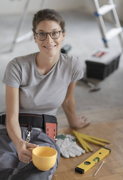 Νέοι χαμογελαστή γυναίκα έχοντας ένα διάλειμμα για καφέ και να κάθεται στο πάτωμα, που κάνει μια ανακαίνιση στο σπίτι - Φωτογραφία, εικόνα
