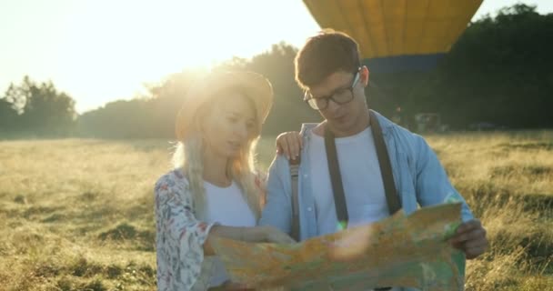 Caucasian young attractive man and woman standing in the field with a map on a sunset. Big balloon on the background. - 映像、動画