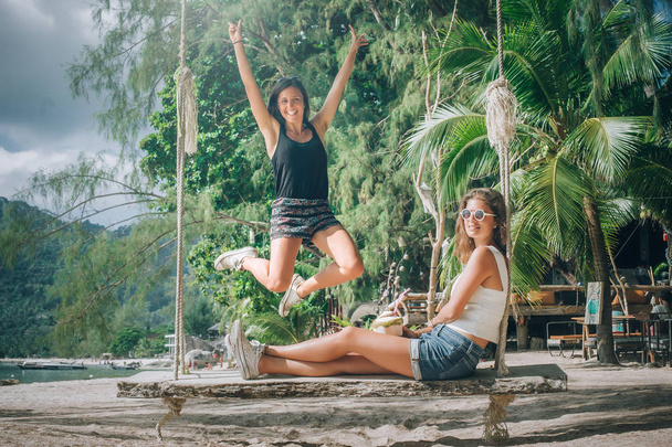 Two beautiful girlfriend enjoy a swing and drink coconut on the beach. Tropical travel holidays - Foto, imagen