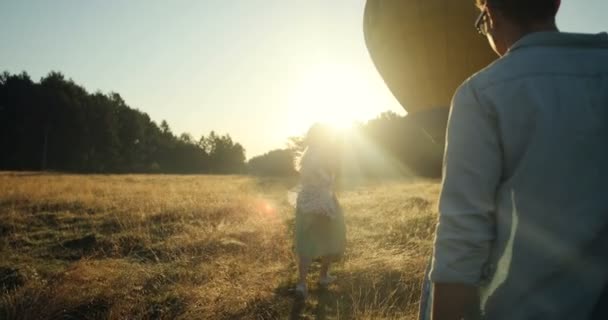 View from behind on the Caucasian young man and woman walking in the field to the big balloon on the picturesque sunset. - Felvétel, videó