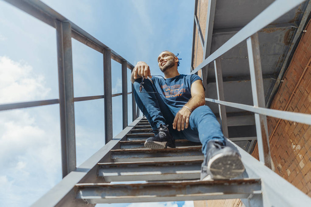 Young attractive man with blue dreadlocks sitting on staircase. Man is on foreground, grey stair is on background. - Foto, Imagen