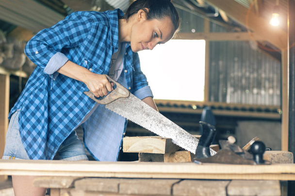 Female carpenter with hacksaw, sawing the boards in the workshop - Photo, Image