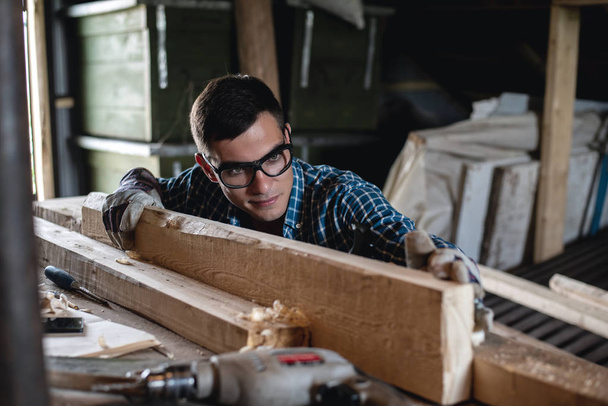 Jovem em óculos de segurança e luvas verifica a qualidade da tábua de madeira aplainada, trabalho manual, carpinteiro
 - Foto, Imagem