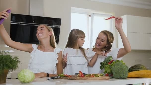 Attractive smiling woman with two daughters do making selfies, photos on smartphone in the kitchen at home. The girls are photographed with vegetables on the smartphone in the home kitchen - Кадри, відео