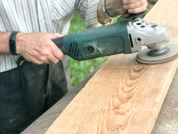El hombre trabaja con sus manos, muele el disco alrededor de la amoladora, la herramienta eléctrica para moler y pulir la superficie de la tabla de madera
 - Foto, Imagen