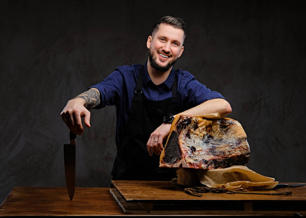 Chef cuisinier souriant tient un couteau et posant près d'une table avec de la viande séchée exclusive sur fond sombre
. - Photo, image