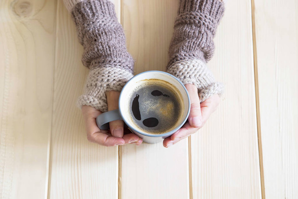 Woman hands with wool warmers holding a cup of coffee on wooden background, top view - Photo, Image