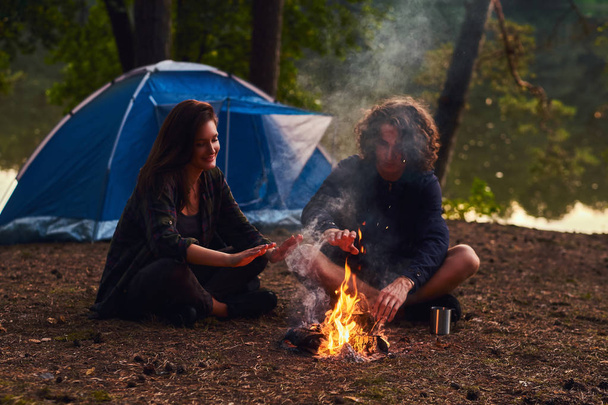 Happy νεαρό ζευγάρι κάθεται και υπερθέρμανση του πλανήτη ένα campfire στο στρατόπεδο στο δάσος κοντά στο ηλιοβασίλεμα. - Φωτογραφία, εικόνα