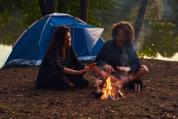 Happy νεαρό ζευγάρι κάθεται και αύξηση της θερμοκρασίας κοντά σε ένα campfire στο στρατόπεδο στο δάσος κατά το ηλιοβασίλεμα. Ταξίδια, τουρισμός και χαράτσι της έννοιας. - Φωτογραφία, εικόνα