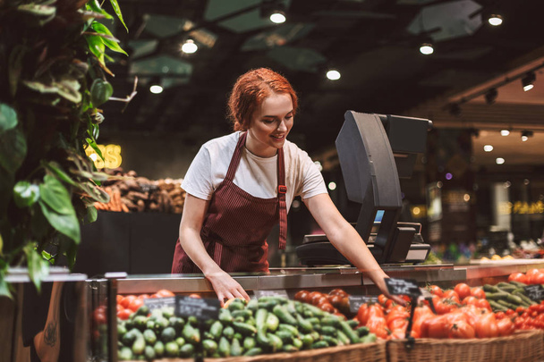 Jonge lachende verkoper in schort staande achter balie met groenten gelukkig werken in moderne supermarkt - Foto, afbeelding
