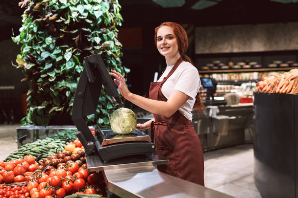 Jonge lachende verkoper in schort achter balie met groenten vreugdevol kijken in de camera weegt kool op schaal in moderne supermarkt - Foto, afbeelding