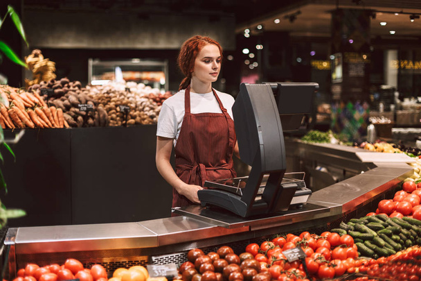 Nuori myyjä esiliina seisoo tiskin takana vihanneksia työskentelee nykyaikaisessa supermarketissa
 - Valokuva, kuva