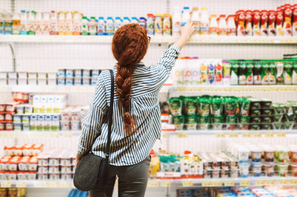 Pretty girl in striped shirt from back choosing dairy products in modern supermarket - Foto, imagen