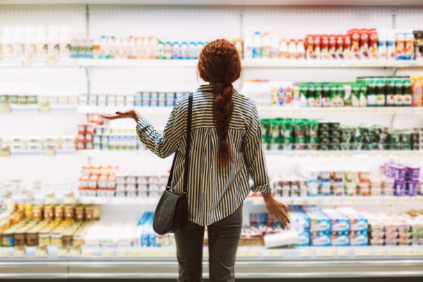 Молодая женщина в полосатой рубашке со спины пытается выбрать молочные продукты в современном супермаркете
 - Фото, изображение