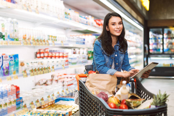 Красивая улыбающаяся девушка в джинсовой куртке с тележкой, полной продуктов, держащих планшет в руке, счастливо смотрящая в камеру в молочном отделе современного супермаркета
 - Фото, изображение