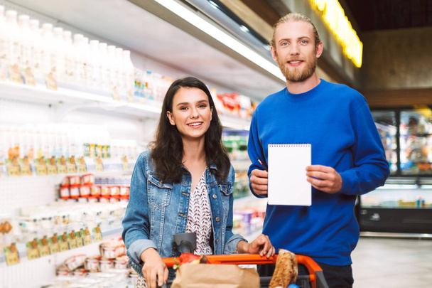 Jeune couple souriant avec liste d'achats et chariot heureux de regarder à la caméra dans le département des produits laitiers du supermarché moderne
 - Photo, image
