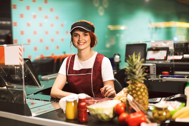 Young beautiful cashier in uniform happily looking in camera working in modern supermarket - Photo, Image