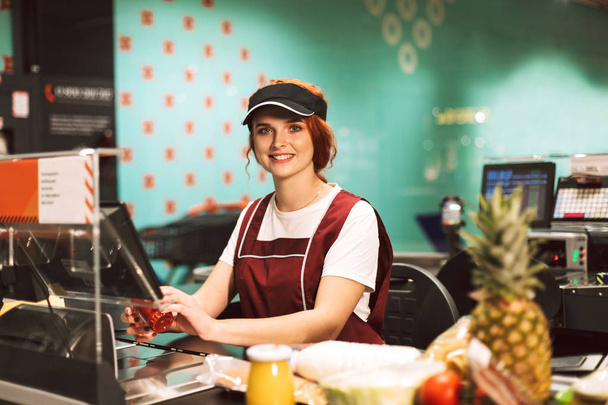 Jovem caixa feminina em uniforme alegremente olhando na câmera trabalhando no supermercado moderno
 - Foto, Imagem