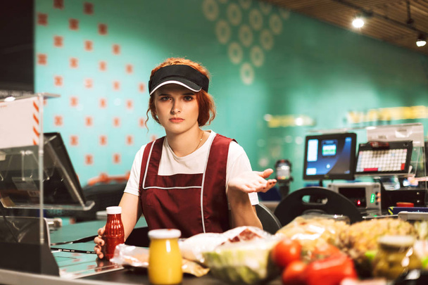 Young upset female cashier in uniform thoughtfully looking in camera working in modern supermarket - Photo, image