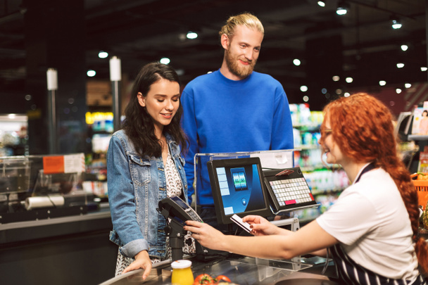 Young smiling couple standing near cashier desk happily paying for products by credit card in modern supermarket - Photo, Image