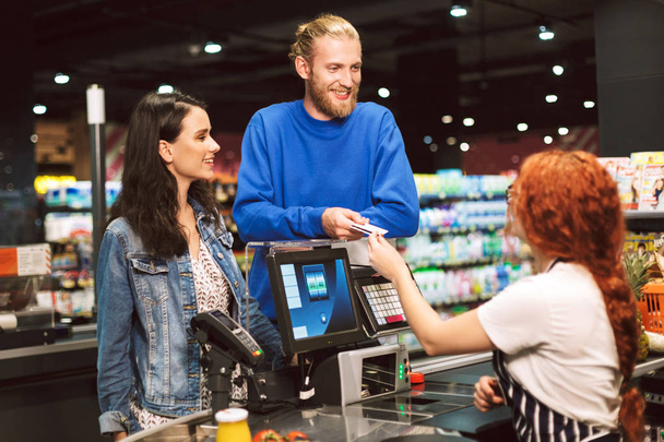 Jovem casal alegre de pé perto da mesa caixa feliz dando cartão de crédito para caixa no supermercado moderno
 - Foto, Imagem