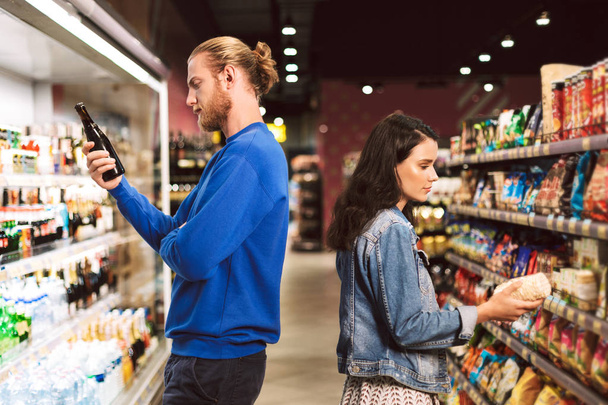 Nuori huomaavainen pari, joka seisoo rinnakkain valitessaan tuotteita, jotka viettävät aikaa nykyaikaisessa supermarketissa
  - Valokuva, kuva