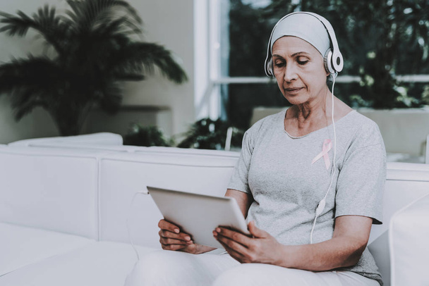 Patient Undergoes Rehabilitation. Cancer Patient on Sofa. Recovering Woman. Remission. Clinic. Cancer Patient. Flowerpots with Flowers. Pink Ribbon. Breast Cancer. Music on Tablet. White Headphones. - Photo, Image