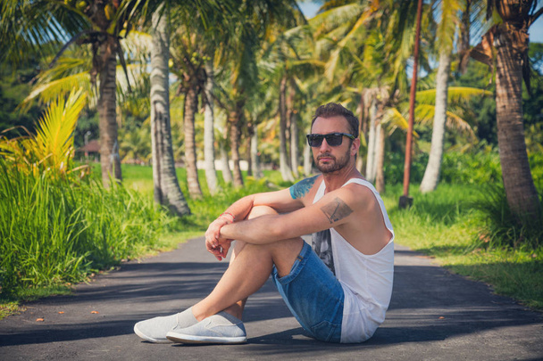 Hombre sentado en medio de un camino vacío con palmeras
. - Foto, imagen