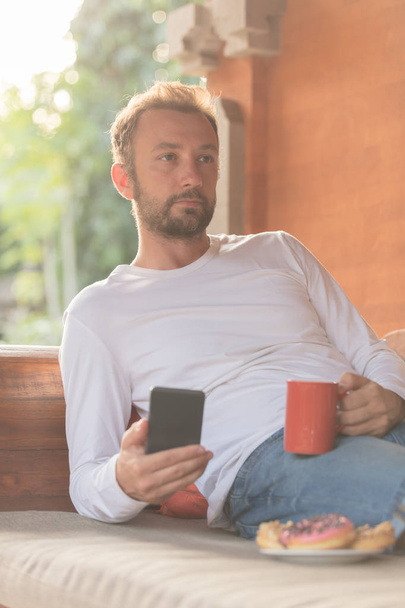 Uomo in possesso di cellulare, tazza di caffè con donazioni sul divano
. - Foto, immagini