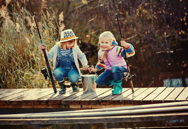 Two little girls with fishing rods, sitting on a wooden pontoon and bragging of caught fish. - Foto, afbeelding