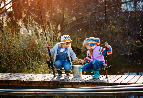Two little girls with fishing rods, sitting on a wooden pontoon and bragging of caught fish. - Foto, afbeelding