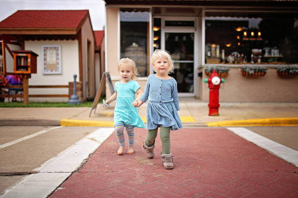 Δύο μικρά κορίτσια νήπια κρατώντας τα χέρια και διασχίζοντας την οδό, ένα το άλλο, οδηγώντας σε μια μικρή πόλη Main Street. - Φωτογραφία, εικόνα
