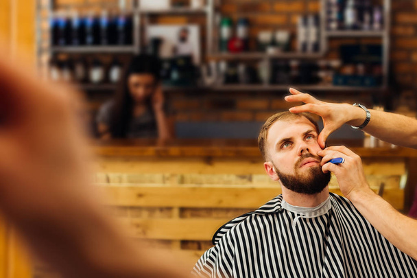 Мужчина в парикмахерской с длинной бородой сидит в кресле, а парикмахер срезает бороду на заднем плане приемной
 - Фото, изображение