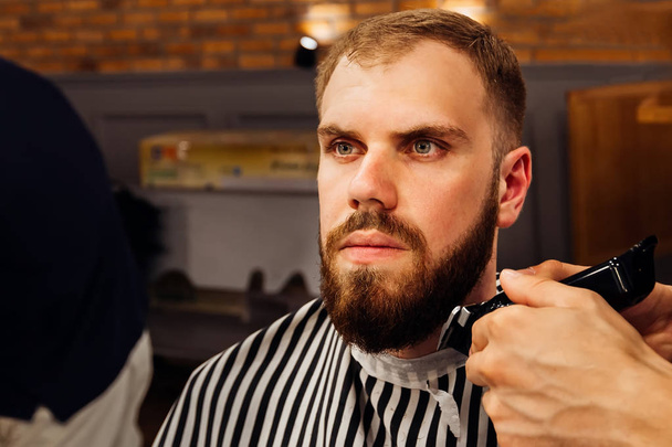a fodrász a trimmer tartja a karját, és teszi egy elegáns és professzionális szakáll, egy ember, egy hosszú szakállal ápolás - Fotó, kép