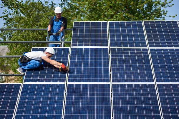 Two technicians working with electrical screwdriver connecting shiny solar photo voltaic panel to metal platform system on green tree thick foliage background. Green energy production concept. - Photo, Image