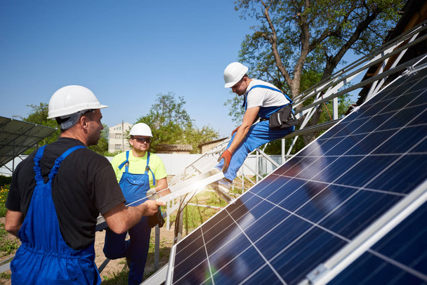 Team von drei professionellen Arbeitern heben schwere Photovoltaik-Solarmodule auf eine hohe Stahlplattform. Solaranlage im Außenbereich, alternatives Konzept zur Erzeugung erneuerbarer grüner Energie. - Foto, Bild