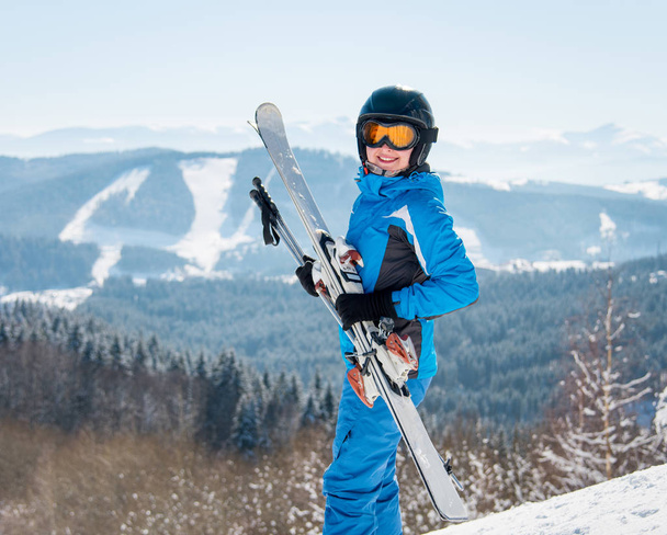 Happy female skier smiling to the camera, holding her skis, wearing blue ski suit and black helmet at winter ski resort happiness positivity vacation travelling - Photo, Image