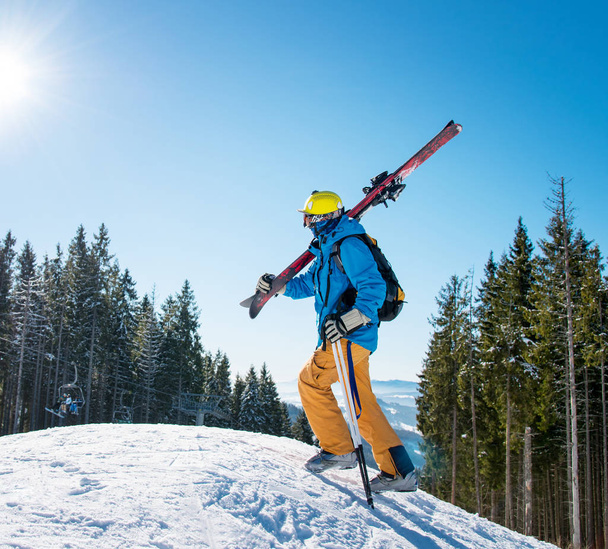 Aufnahme eines männlichen Skifahrers, der an einem sonnigen Tag in einem Wintersportort mit aktivem Lifestyleausrüstungskonzept den Hang in den Bergen hinaufgeht und seine Ausrüstung auf der Schulter trägt.. - Foto, Bild