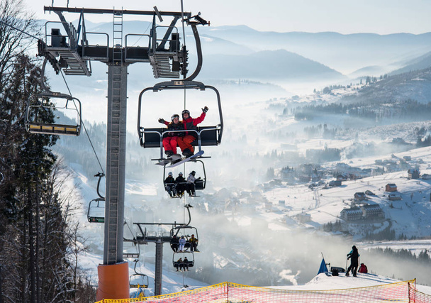 Snowboarders y esquiadores en un telesilla en la estación de esquí de invierno con un hermoso fondo de pistas cubiertas de nieve, bosques, colinas. Bukovel, Ucrania
 - Foto, Imagen