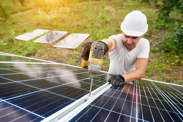 Smiling technician working with screwdriver connecting solar photo voltaic panel to exterior metal platform on sunny green orchard and rural houses background. Efficiency and professionalism concept. - Photo, Image