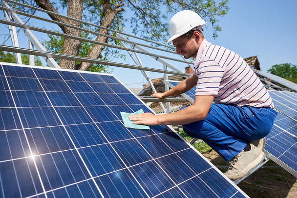 Profile of male worker in white helmet wiping clean big square shiny solar panel standing on tall metal platform on summer bright blue sky background. Renewable energy generation concept. - Valokuva, kuva