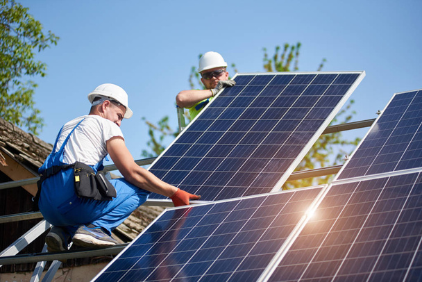 Dos técnicos profesionales que ajustan el panel fotovoltaico solar pesado a la plataforma del metal en el fondo azul del cielo. Instalación, eficiencia y profesionalidad del sistema de paneles solares autónomos
. - Foto, imagen
