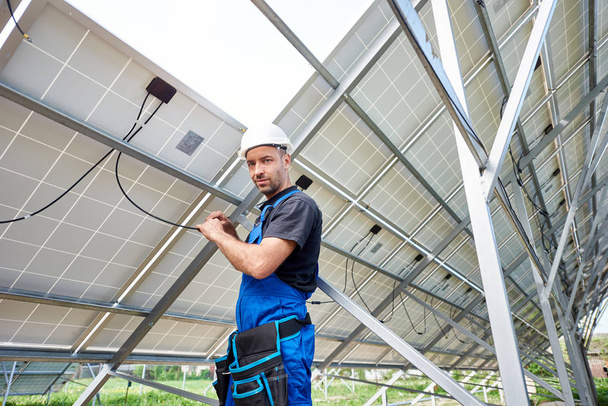 Jeune technicien ingénieur faisant le câblage électrique debout à l'intérieur du système photovoltaïque extérieur haut panneau solaire, regardant vers la caméra. Eco friendly concept de production d'électricité bon marché
. - Photo, image