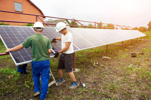 Команда робітників завершує збірку зовнішньої автономної сонячної фотовольтаїчної панелі в зеленому полі перед сільським котеджем в сонячний літній день. Альтернативна відновлювана екологічна зелена енергія
. - Фото, зображення