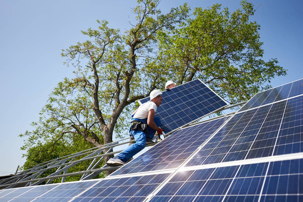 zwei Arbeiter mit Schutzhelm auf einer hohen Stahlplattform, die schwere Solar-Photovoltaik-Panels auf grünem Baum und blauem Himmelshintergrund montiert. Solarpaneel-Installation im Außenbereich, gefährliches Jobkonzept. - Foto, Bild