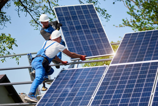 zwei Arbeiter montieren schwere Solar-Photovoltaik-Panel auf einer hohen Stahlplattform auf grünem Baum und blauem Himmel Hintergrund. Stand-Alone-Solarpaneelsystem für den Außenbereich, gefährliches Arbeitsplatzkonzept. - Foto, Bild