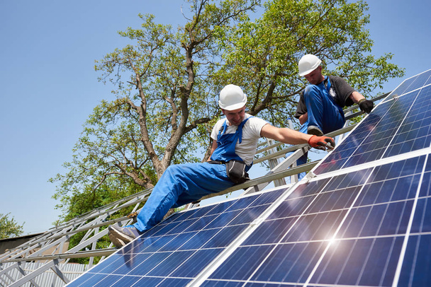 Two technicians on high metal platform against bright blue sky and green tree working on voltaic solar panel system installation. Renewable ecological cheap green energy production concept. - Photo, Image