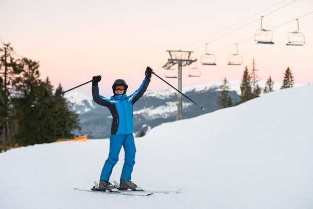 Girl enjoying ski holiday standing on the snowy mountain and raised her hands up. Woman at ski resort wearing helmet, blue ski suit and goggles. - Photo, Image
